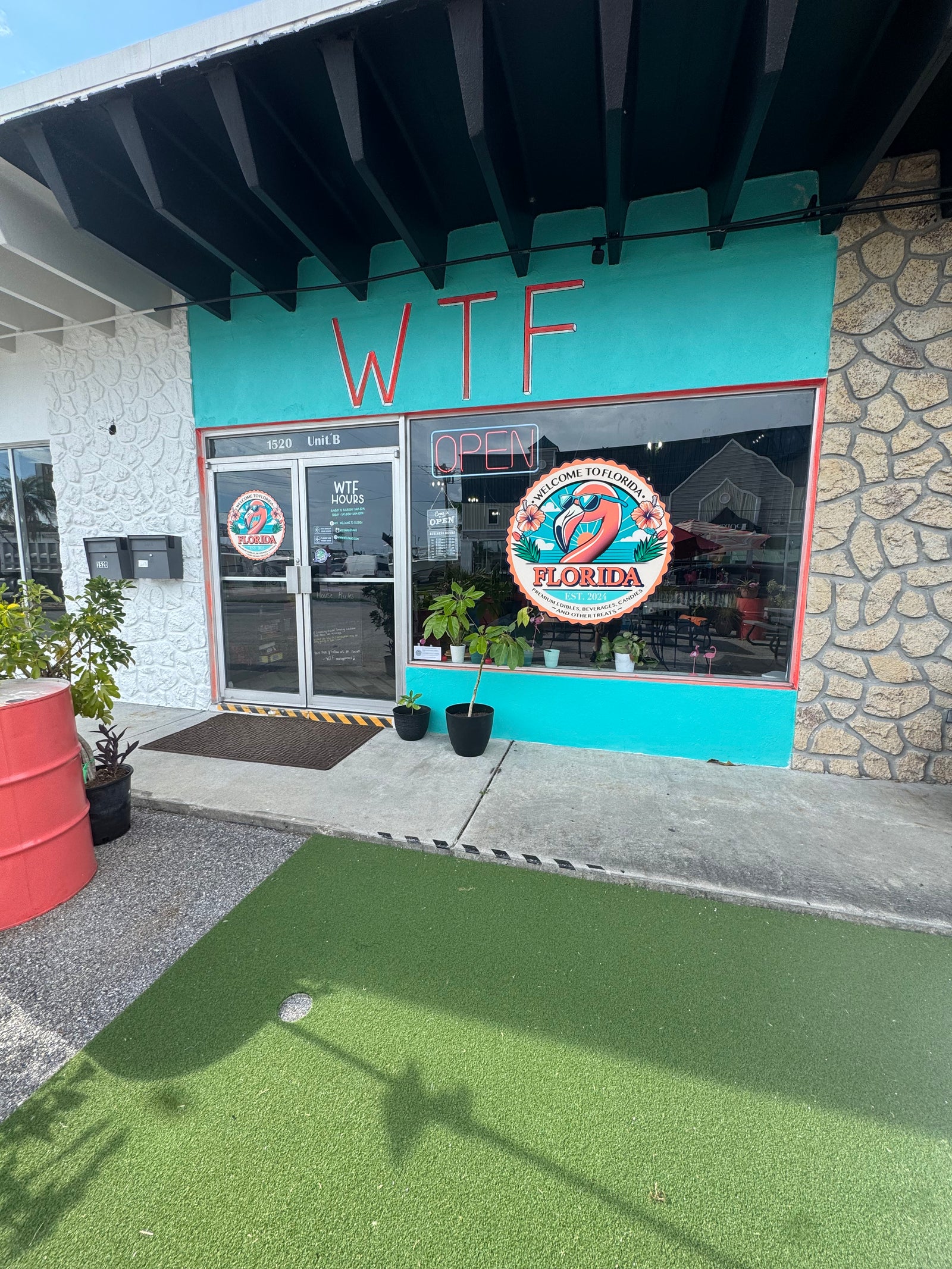 The storefront of WTF in Florida, featuring a bright teal facade, a 'Welcome to Florida' flamingo sign in the window, and a mini-golf putting green out front.